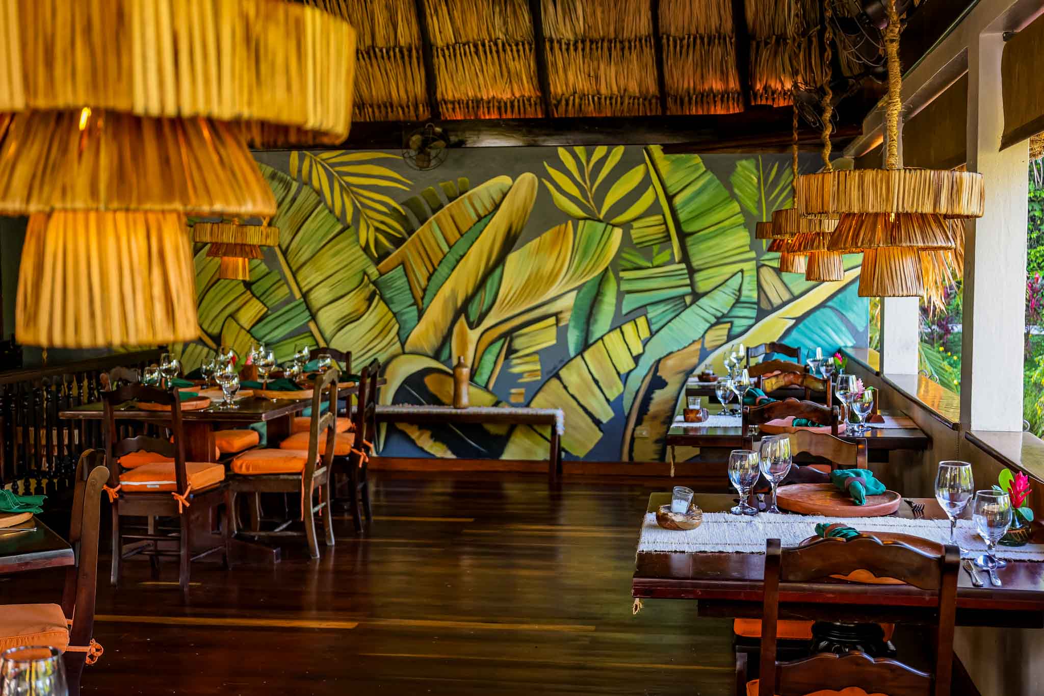 Comedor del Restaurante Grove House con un vibrante mural de hojas de plátano en la pared, luminarias con flecos de paja y mesas de madera preparadas